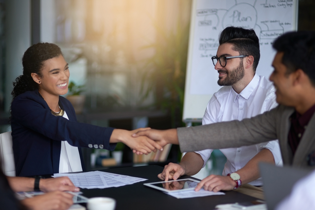 Two professionals shaking hands across a table during a customer relationship management meeting, with colleagues discussing in the background.