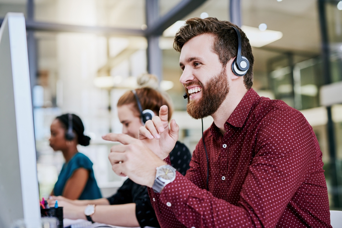 A smiling man wearing a headset gestures at a computer screen displaying customer relationship management software in a busy office setting with colleagues around him.