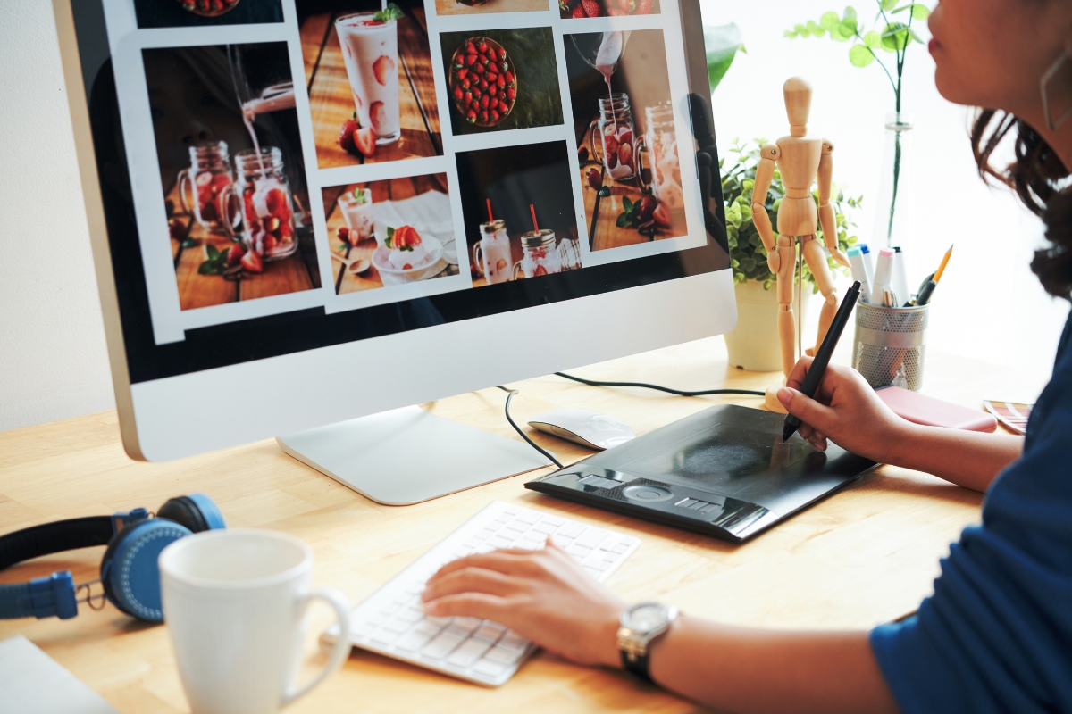 Graphic designer using a stylus and tablet to edit photos of beverages on a computer screen with Divi Builder themes in a bright office space.