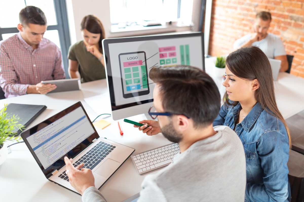 Two professionals discussing a mobile app design using Divi Builder themes on a computer screen in a modern office setting, with colleagues working in the background.