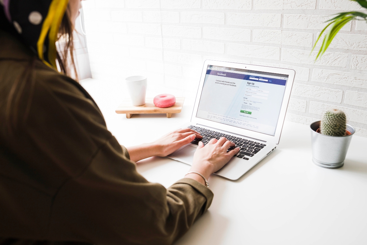 Woman using a laptop to analyze a Facebook marketing mistake on a white desk with a coffee mug and a small cactus nearby, in a well-lit room.