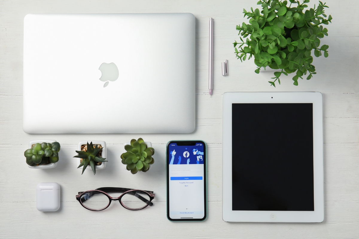 A neatly organized workspace featuring a laptop, tablet, smartphone, eyeglasses, potted plants, and stationary items on a white wooden surface dedicated to Facebook marketing strategies.