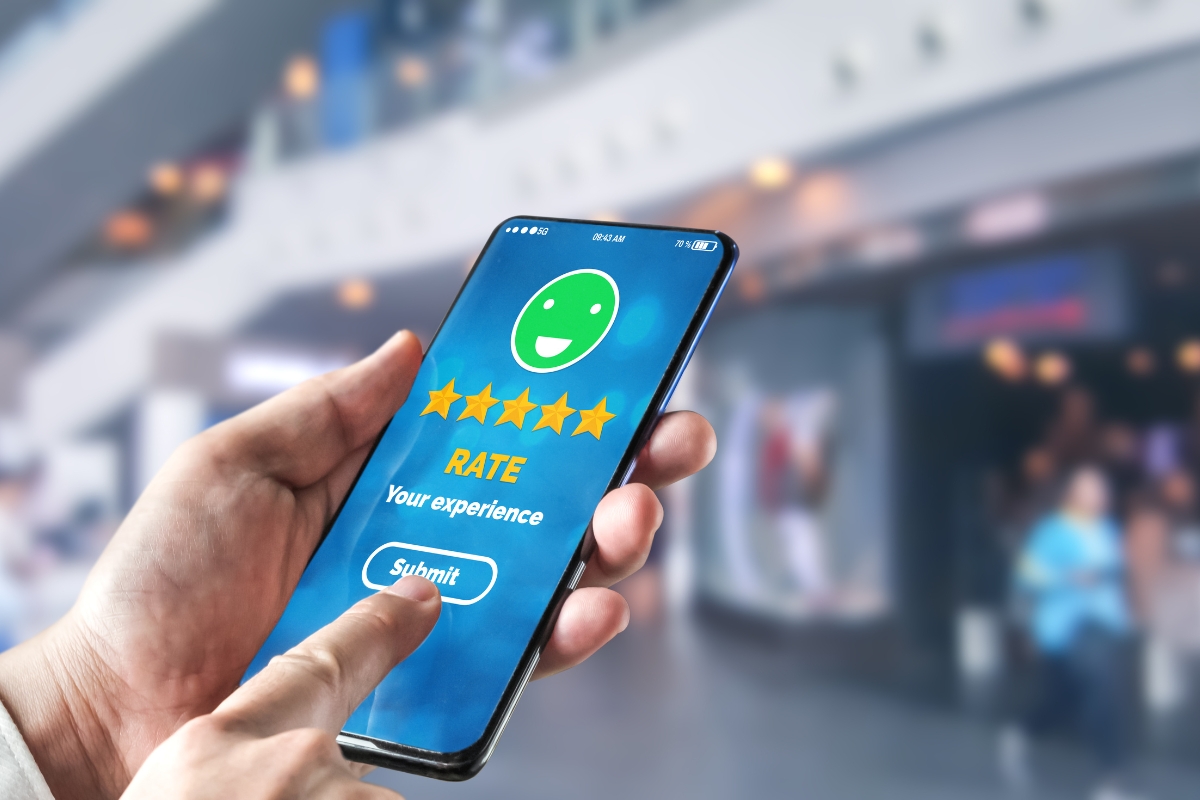 A person's hand holding a smartphone displaying Google reviews with a five-star rating and a green happy face, in a busy public space.