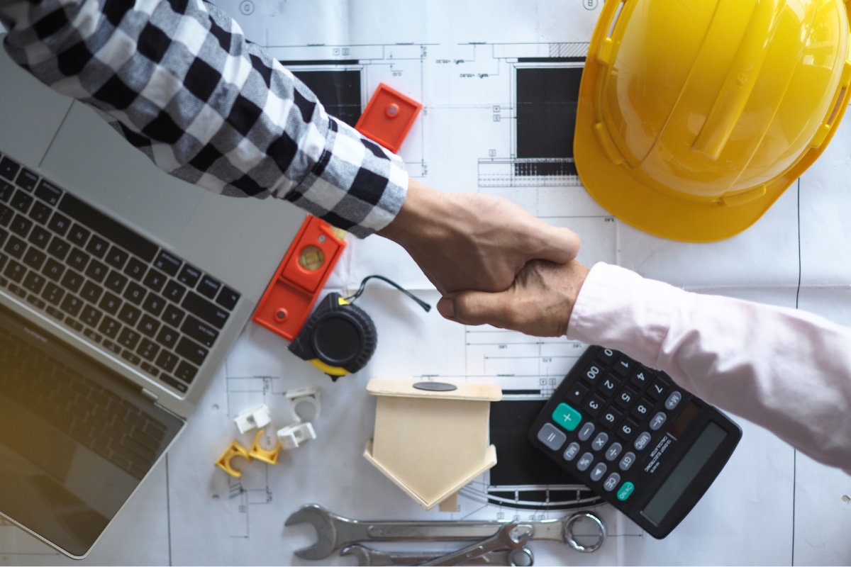 Two people shaking hands over a desk with construction plans, a laptop, hard hats, and engineering tools on Houzz Pro.