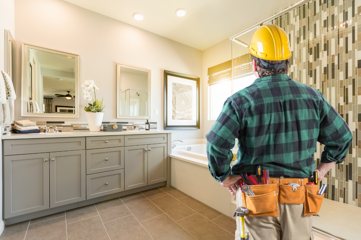 A construction worker wearing a yellow helmet and a tool belt stands facing a modern bathroom with a dual vanity and decorative tiling, designed using Houzz Pro.