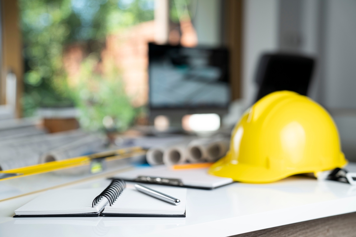 A focused image depicting a construction helmet, notebook, and a mobile phone on a desk with blurry architectural plans and a laptop in the background, all part of a Houzz Pro setup.