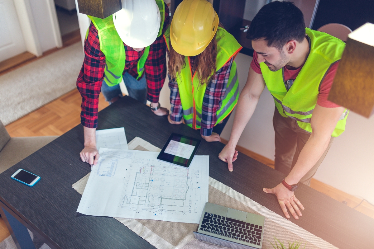 Three construction workers in hard hats and safety vests examining blueprints on a table with a laptop and smartphone nearby, utilizing Houzz Pro for project management.