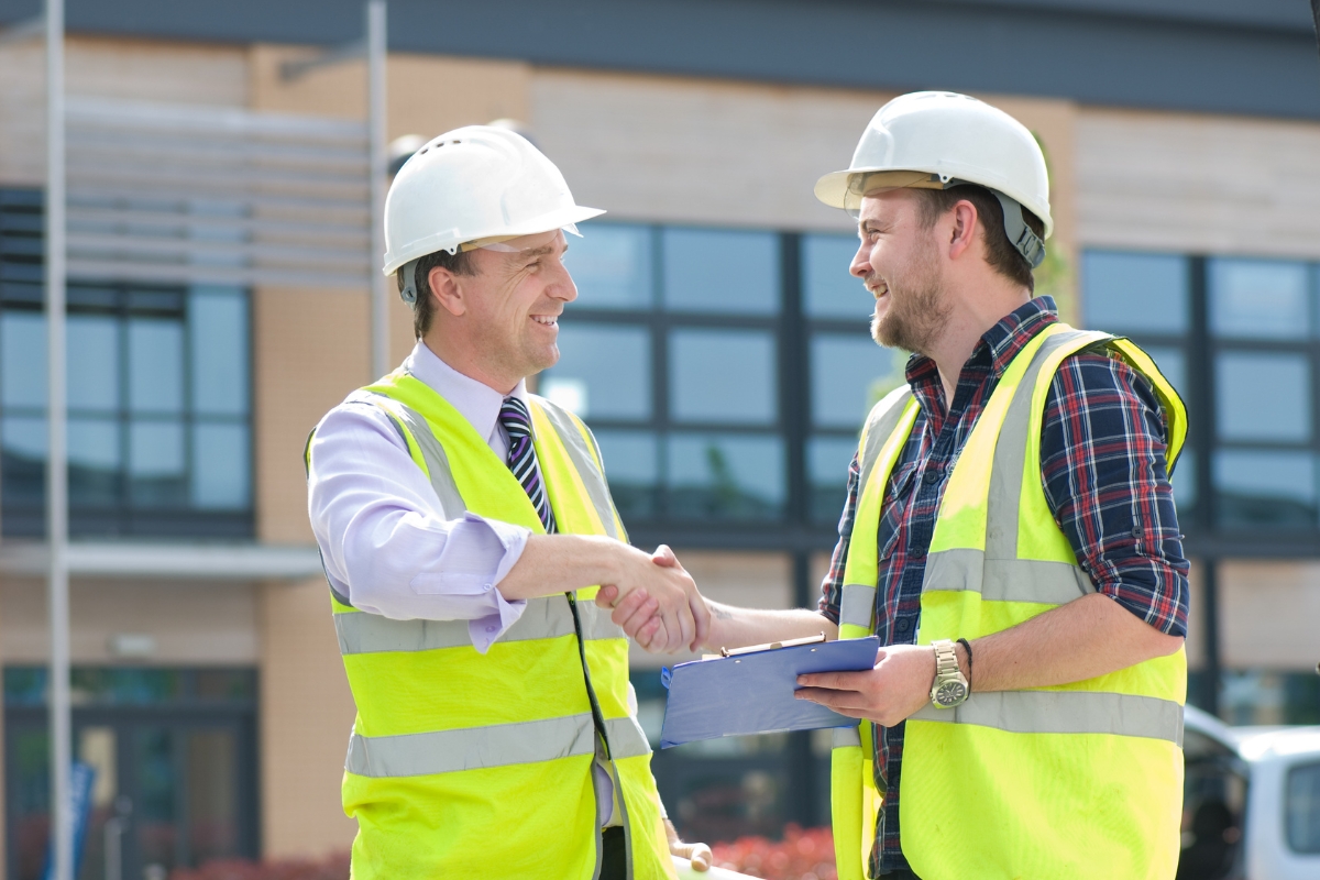 Two construction workers wearing hard hats and high-visibility vests shaking hands in front of a building on Houzz Pro.