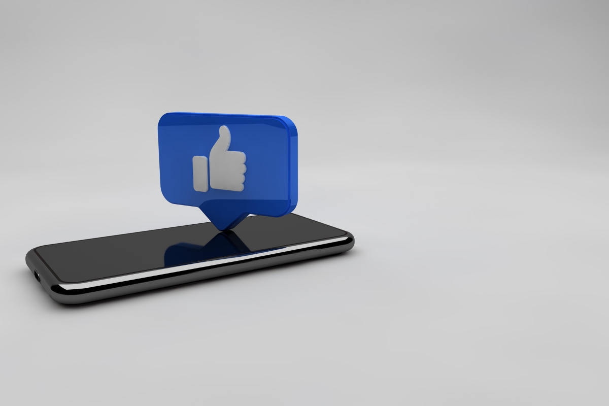 A digital rendering of a 3d blue "like" icon emerging from a smartphone screen against a white background, symbolizing how to improve your Facebook business page.