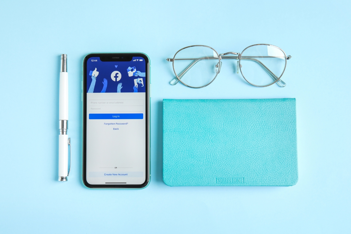 Smartphone displaying how to improve your Facebook business page, placed next to a pen, eyeglasses, and a teal notebook on a light blue background.