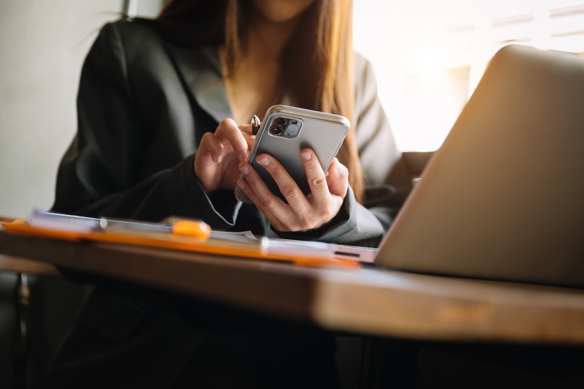 A woman in a blazer using a smartphone to improve her Facebook business page, next to her laptop at a desk with documents under warm lighting.