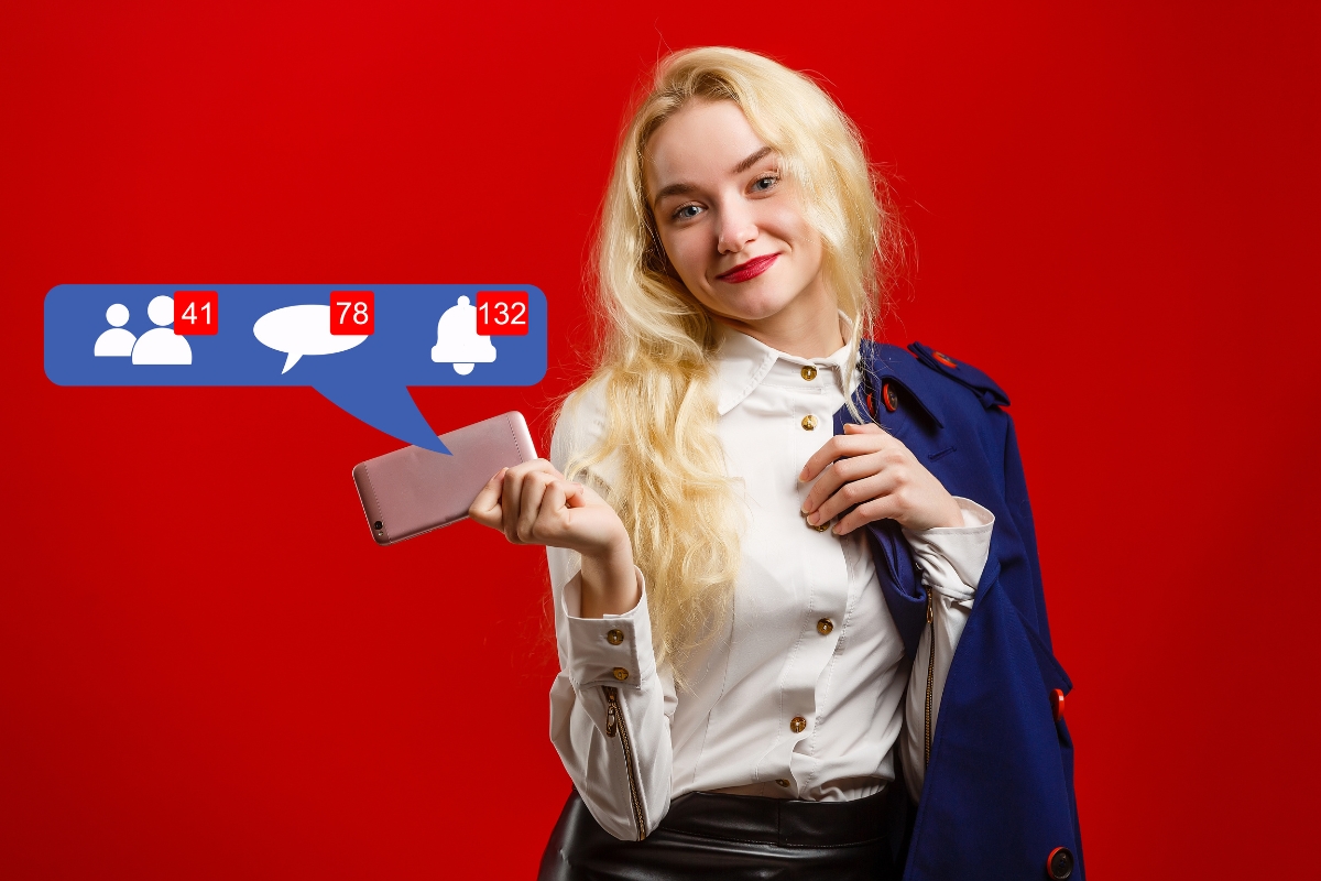 A woman with blonde hair smiles while holding a smartphone displaying how to improve your Facebook business page; she wears a white blouse and a navy jacket against a red background.