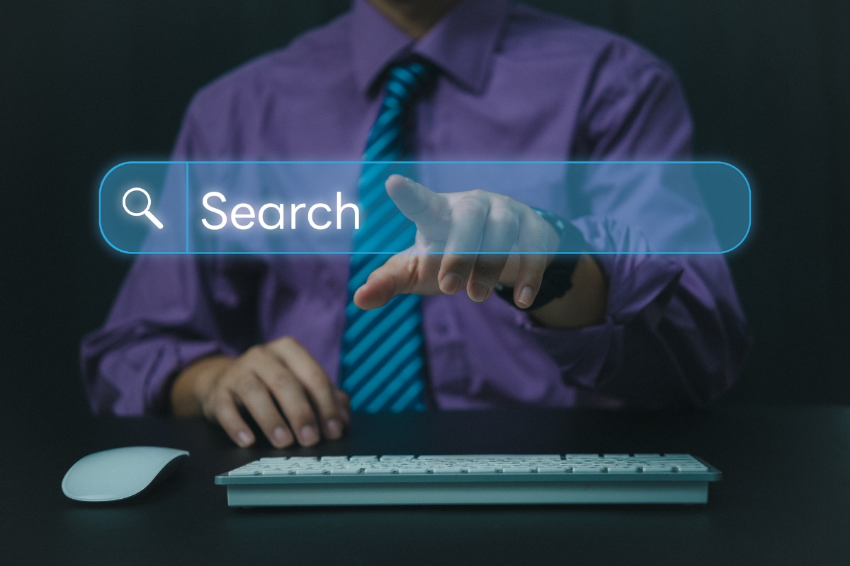 A man in a purple shirt and tie sitting at a desk, pointing at a digital search bar projected in front of him, demonstrating how to search on a website.