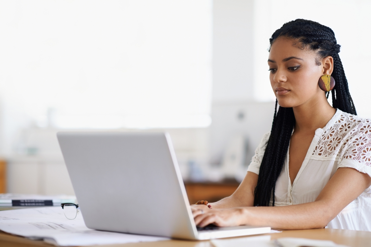 A focused young woman with braided hair learning how to search on a website using a laptop at a desk in a brightly lit office.