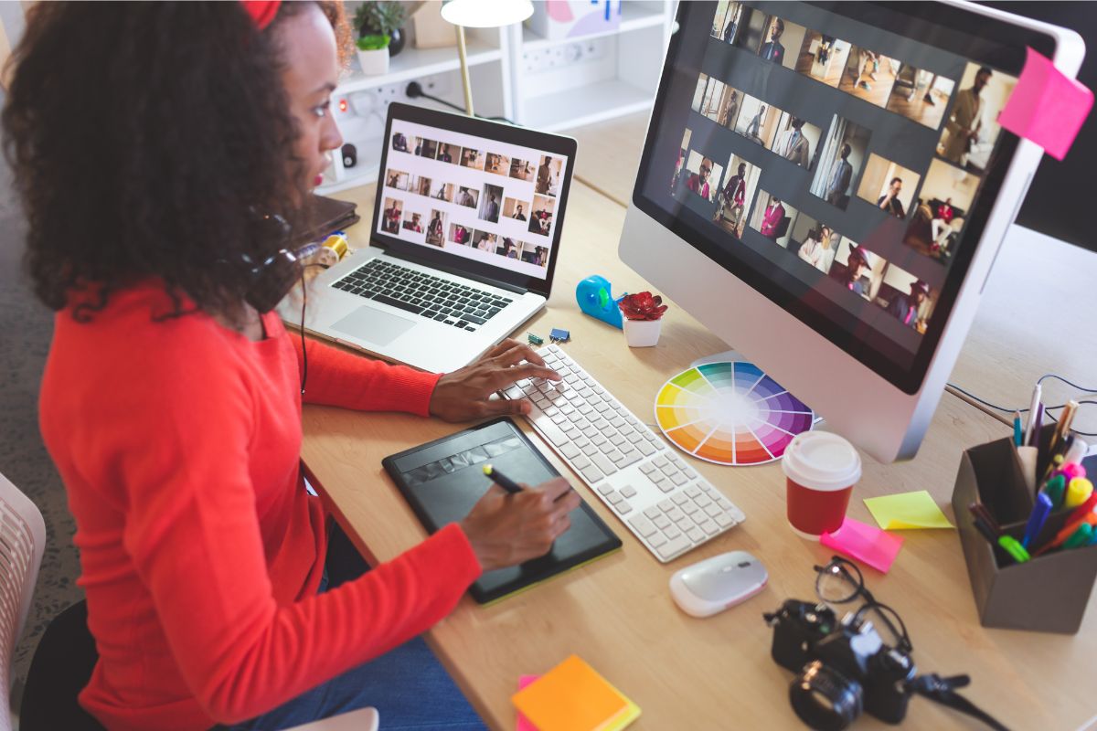 A woman in a red sweater using a stylus on a tablet at a desk, with a computer screen displaying multiple video call participants and images for SEO.