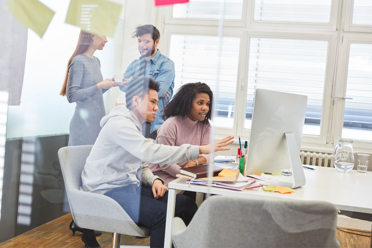 Four colleagues collaborating at a desk with computers, papers, and images for SEO in a bright office space. Two are discussing while the others are engaged in tasks.