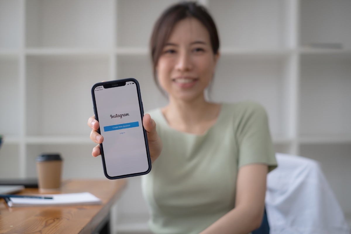 A woman in a light green shirt smiling and holding a smartphone showing the Instagram marketing tips screen, with a blurred coffee cup and light interior background.