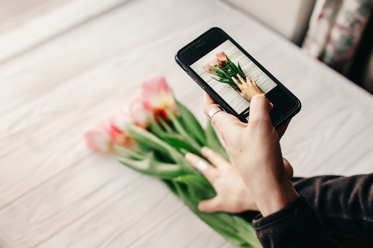 A person's hand holding a smartphone taking a photo of pink tulips on a wooden table for Instagram marketing tips.