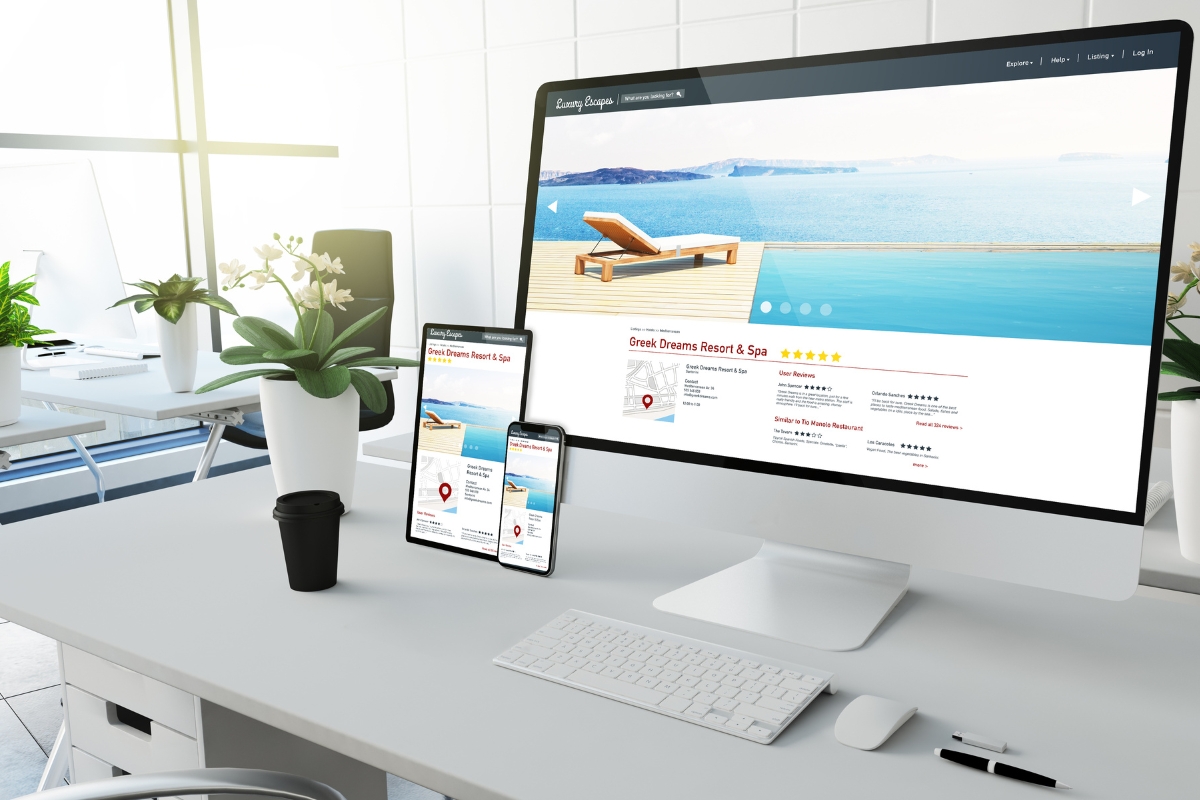 Modern workspace with a desktop computer, tablet, and smartphone displaying the same travel website featuring local business citations, a cup of coffee, and a potted plant on a white desk.