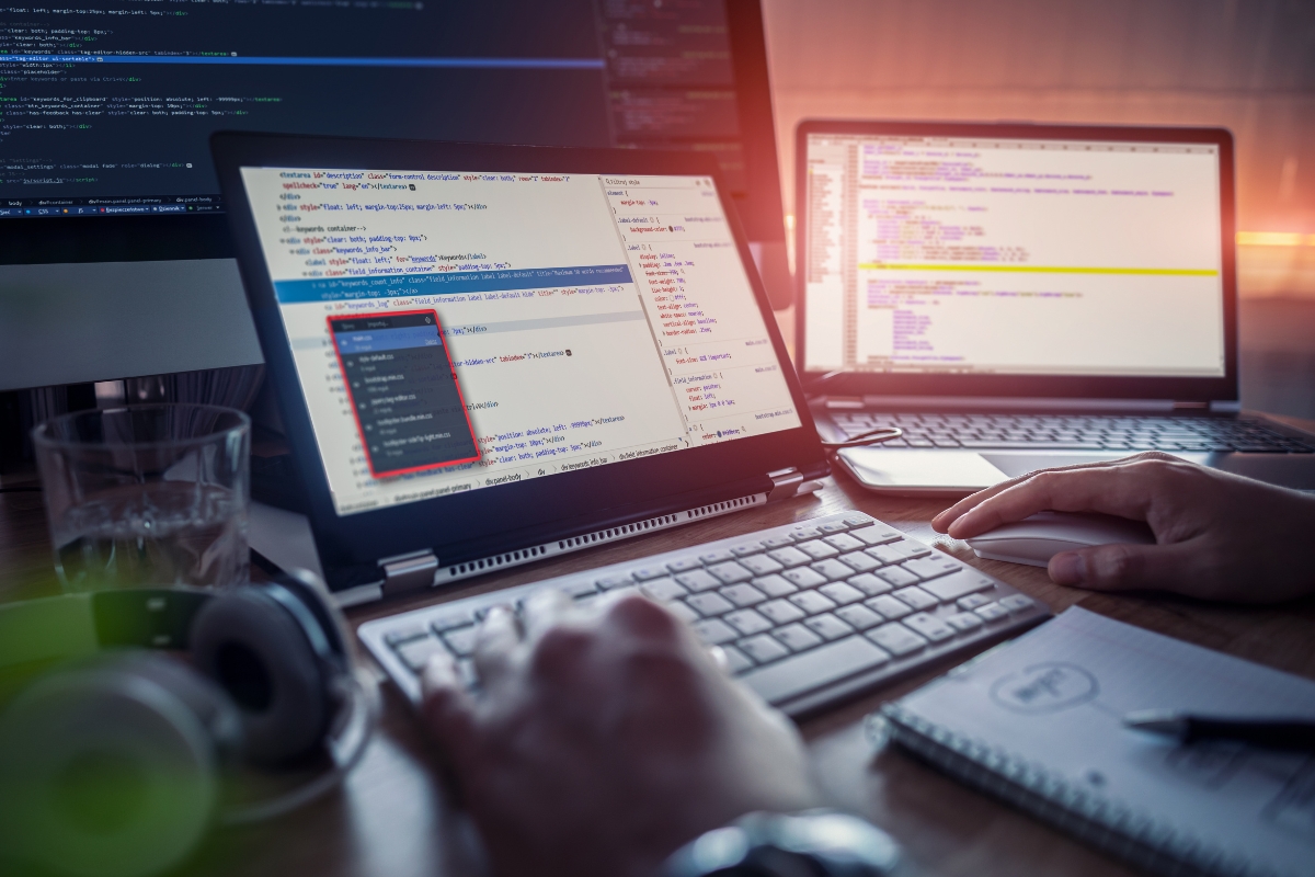 A person managing WordPress sites on multiple monitors displaying code, in a dimly lit room with visible accessories like a notebook, headphones, and a smartphone.