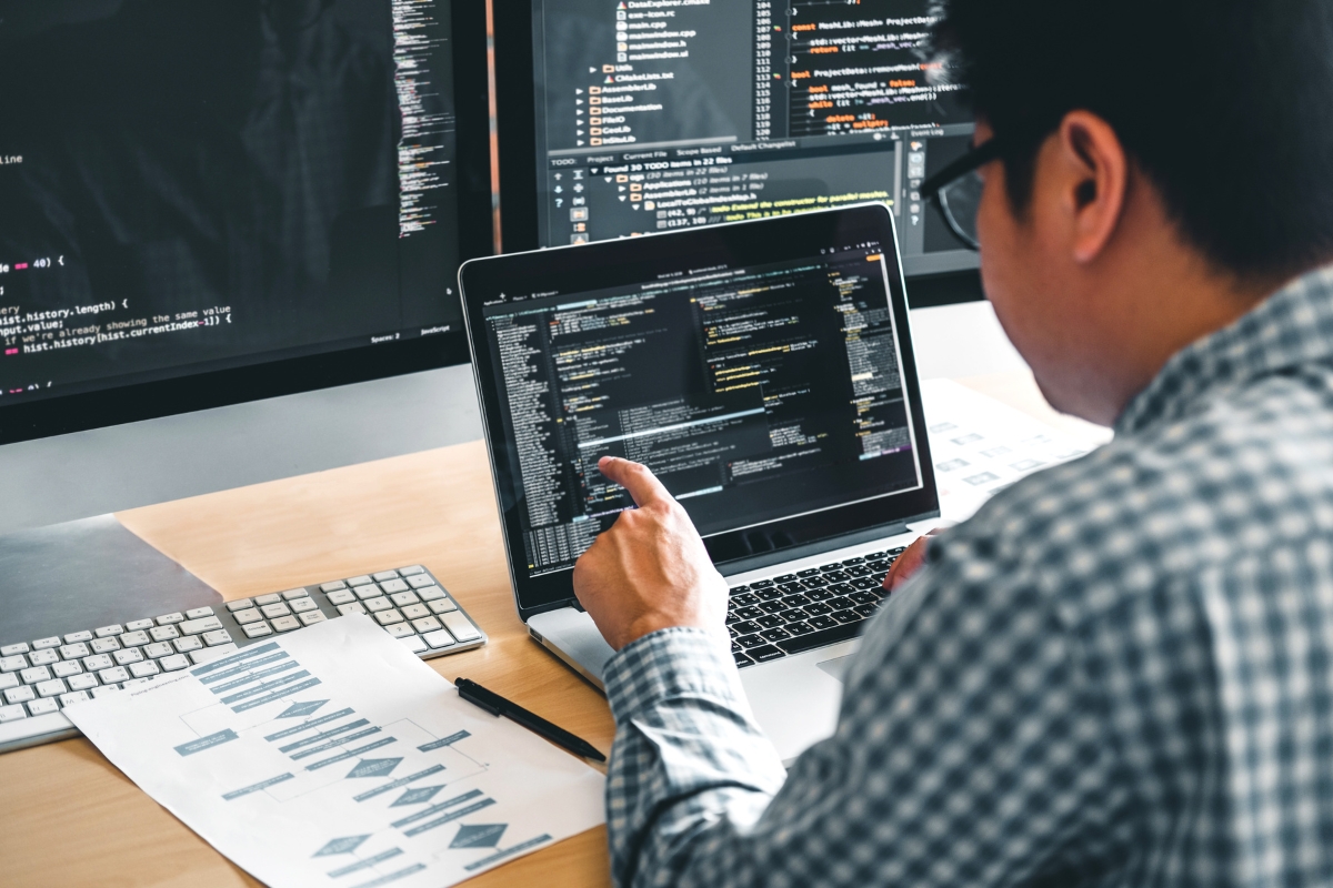 A man in a plaid shirt managing WordPress sites on a laptop with code on the screen, papers and a second monitor visible on a desk.