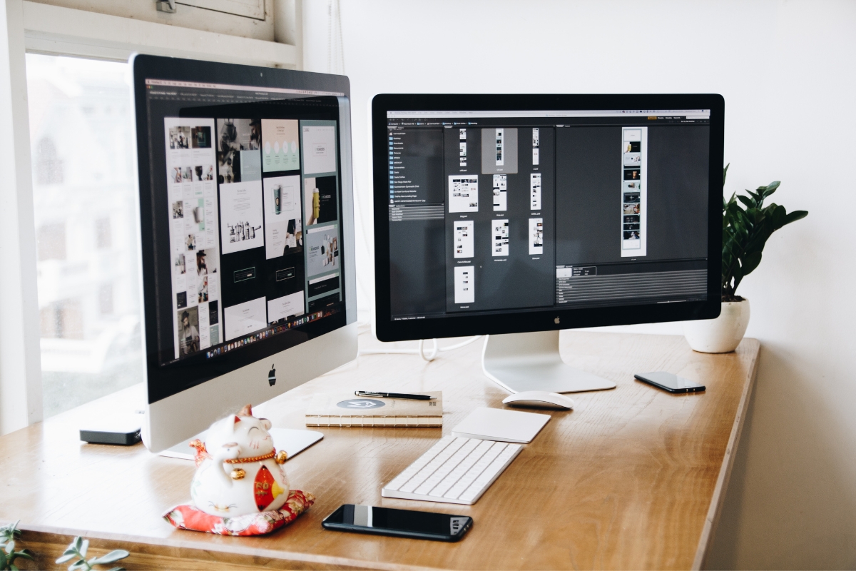 Two computer monitors on a desk displaying managing WordPress sites software, with a phone, a small plant, and a decorative figurine alongside.