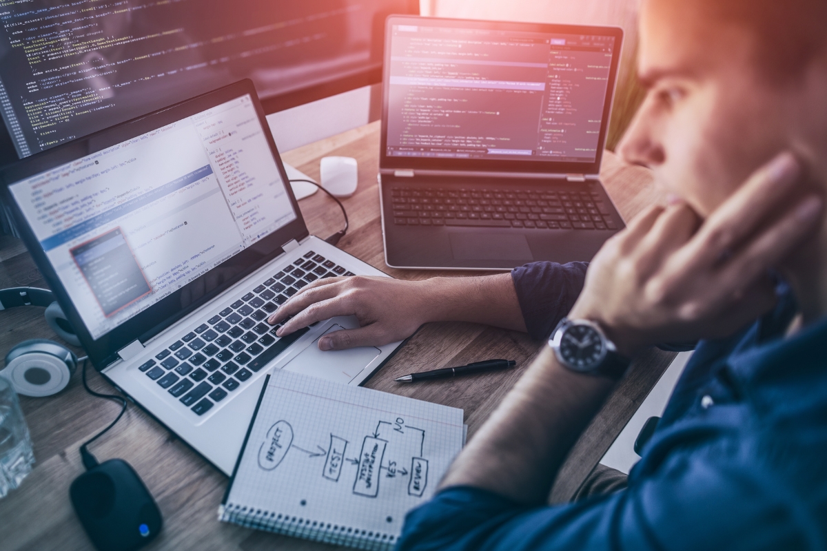 Male programmer managing WordPress sites on a laptop with multiple screens displaying code, sitting at a desk with notes and a coffee cup.