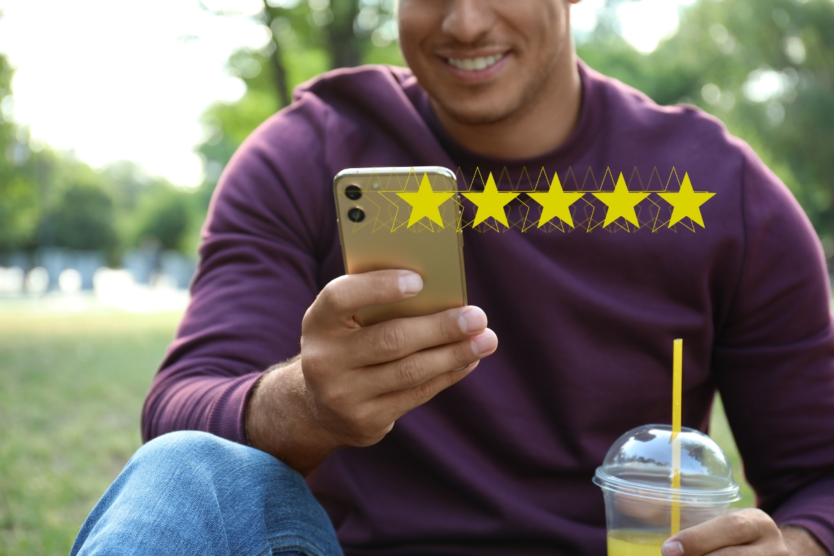 Man in a purple sweater sitting in a park, holding a smartphone displaying five gold stars indicative of his positive online reputation, with a cold drink beside him.