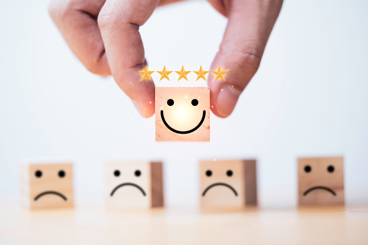 Hand holding a wooden block with a smiley face and five stars above it, representing a positive online reputation, standing out among blocks with sad faces.