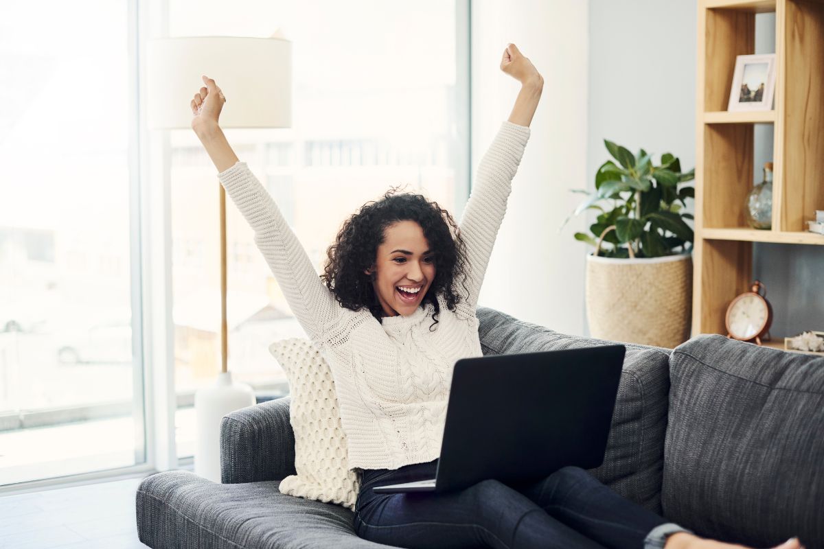 A joyful woman with curly hair raises her arms in excitement while looking at a laptop displaying Rank Math SEO on a gray sofa.