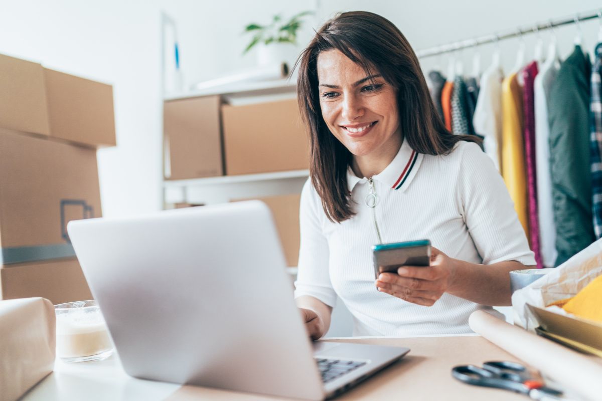 A woman smiles as she uses her smartphone and laptop in a home office with sewing materials and clothes in the background, researching how to create a Facebook business account.