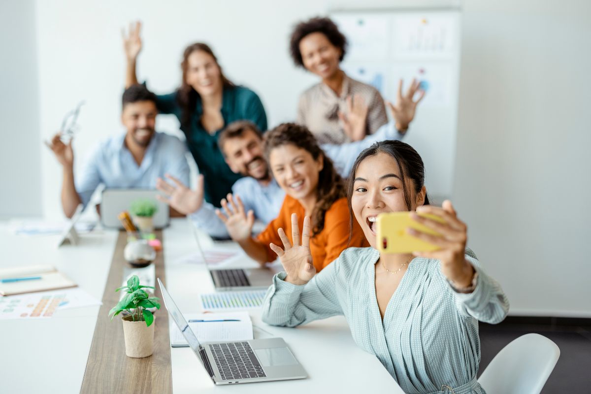 A diverse group of six colleagues in a bright office taking a selfie for employee advocacy on social media, smiling and waving at the camera, with laptops and documents on the table.