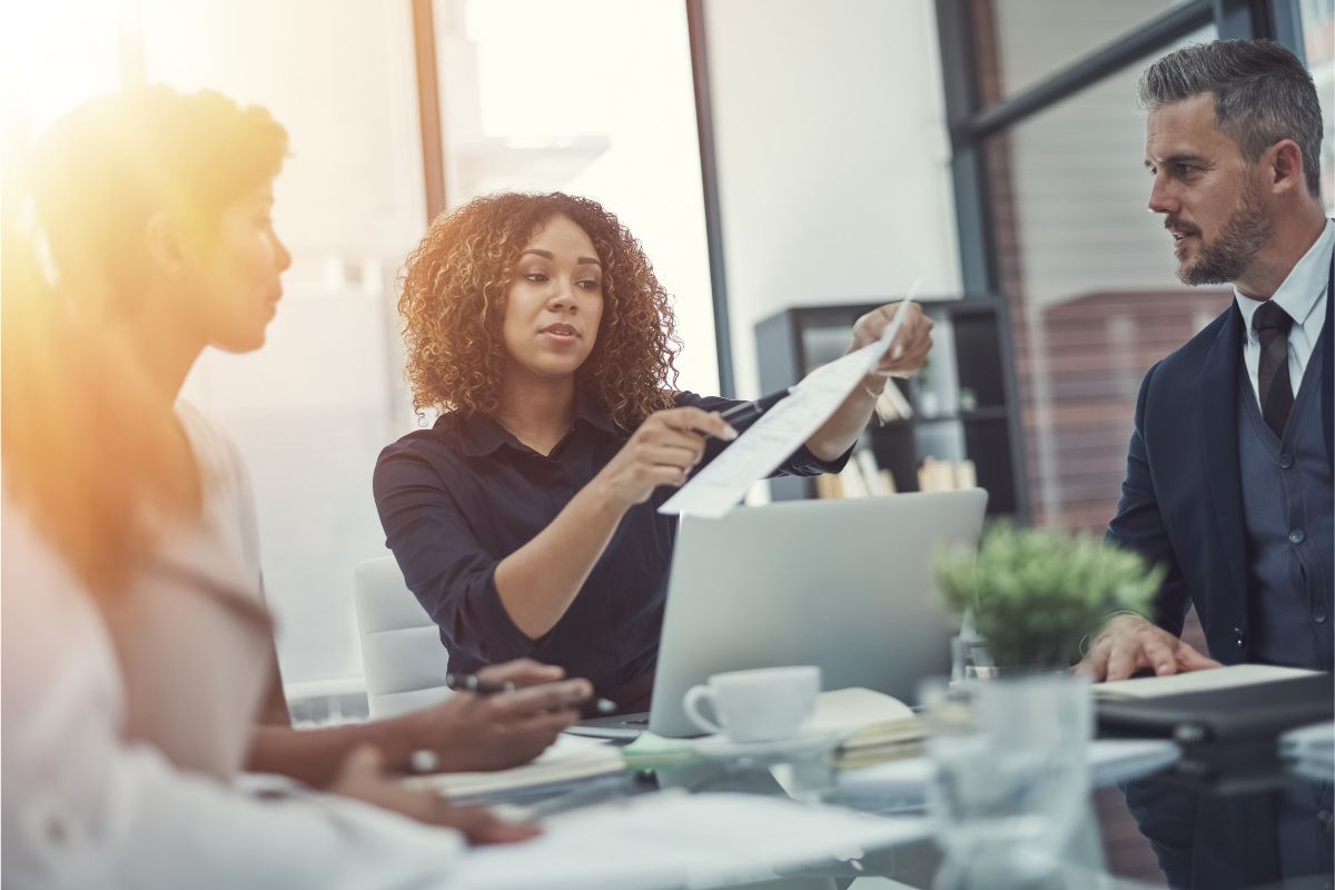 Three people in a business meeting. A woman in the center is showing a document to a man on the right, explaining how to use ChatGPT effectively. Another woman on the left listens attentively.