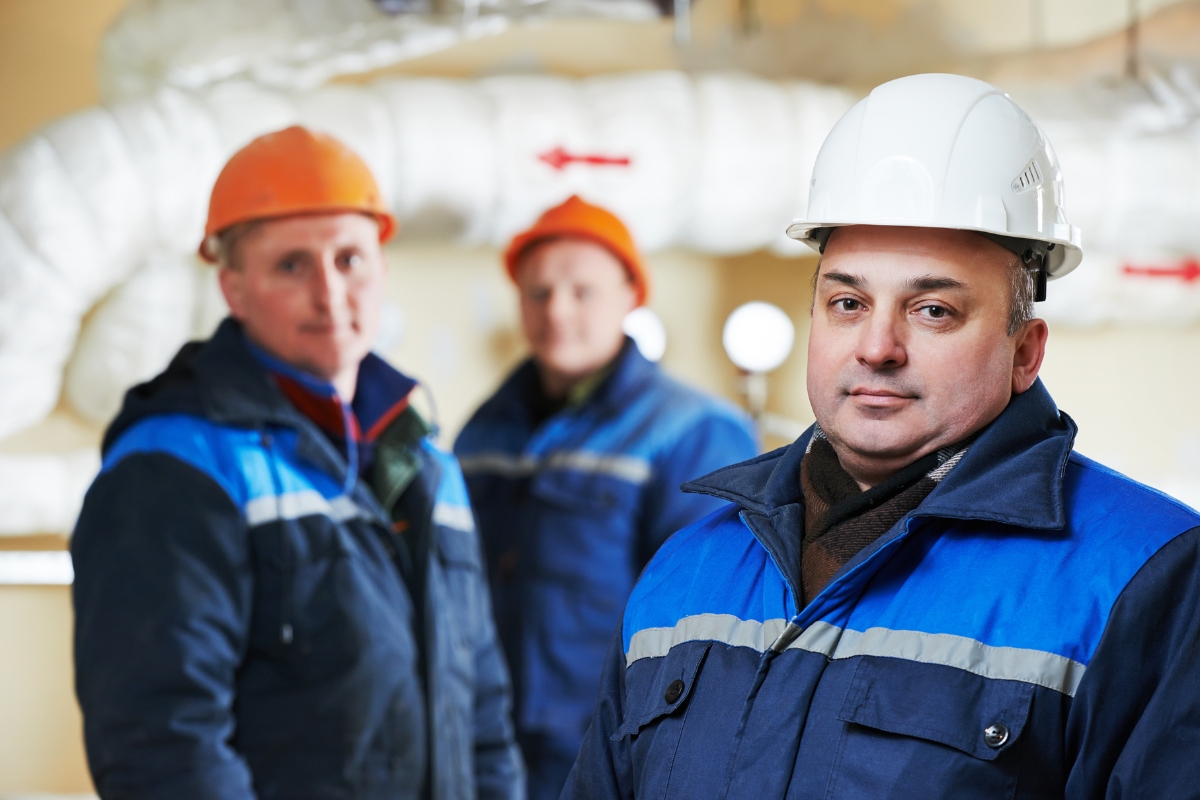 Three men wearing hard hats and blue work jackets stand indoors with industrial pipes in the background. The man in the foreground, representing community marketing, is in sharp focus, while the other two are slightly blurred.