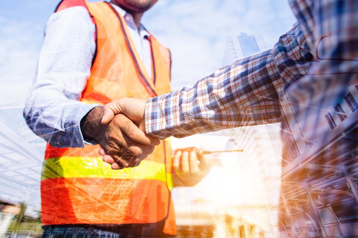 Two people wearing safety vests and checkered shirts are shaking hands at a construction site with a cityscape in the background, exemplifying the spirit of community marketing through collaboration and shared goals.