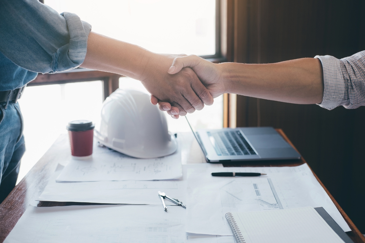 Two people shake hands over a desk with blueprints, a laptop, a coffee cup, and a hard hat—a perfect example of community marketing in action.