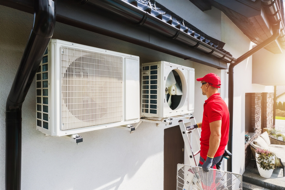 A worker in a red uniform is servicing air conditioning units mounted on the exterior wall of a building. While holding a panel and standing on a ladder, he exemplifies dedication and skill, reflecting the community marketing values of trust and reliability.