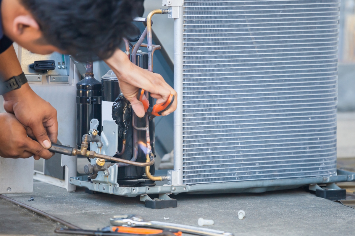 A technician uses a blowtorch to repair or install copper tubing on an air conditioning unit, promoting community awareness through exceptional service. Various tools lie on the ground nearby.