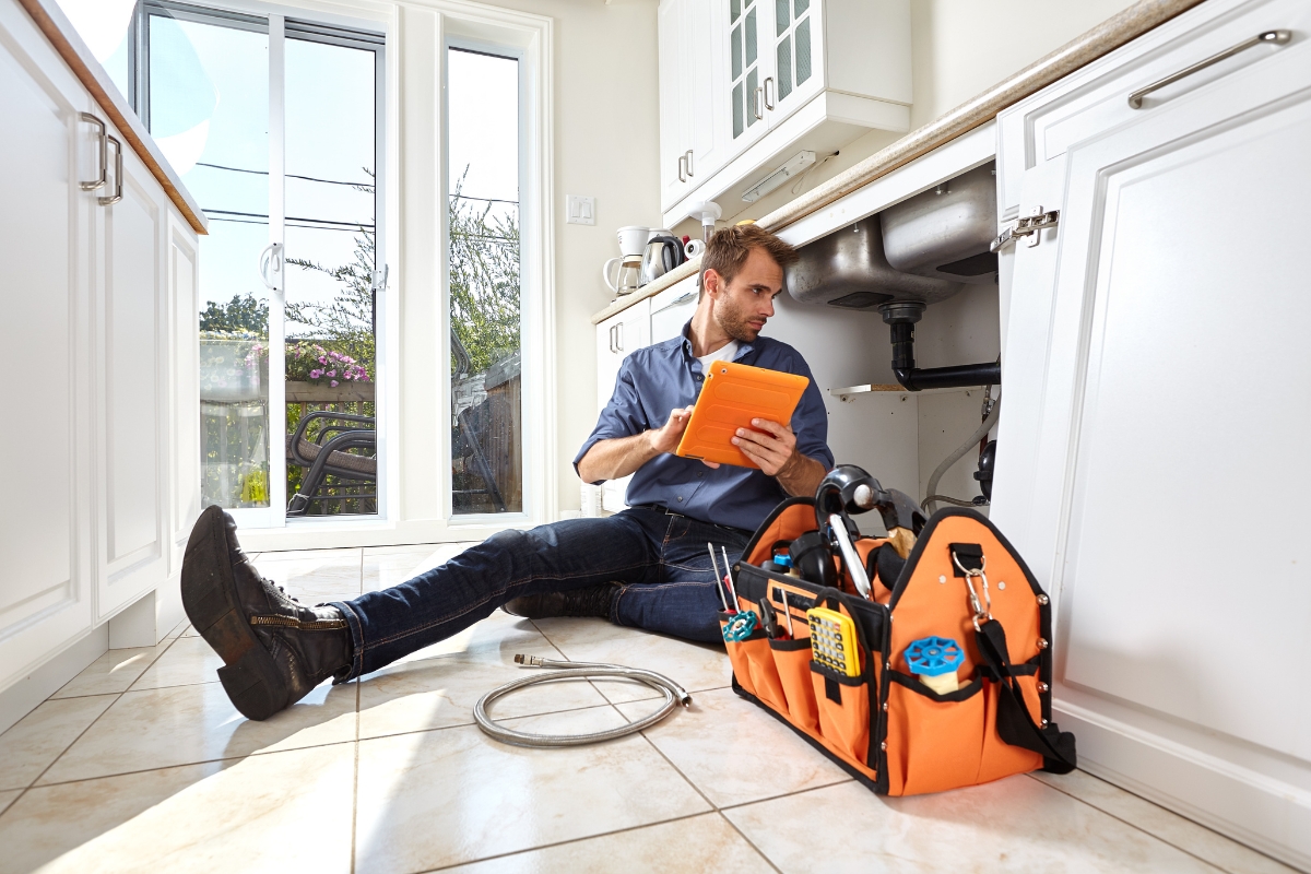 A person is sitting on the kitchen floor with tools, looking at an orange tablet next to an orange tool bag, while working under a sink, possibly researching content marketing for plumbers.