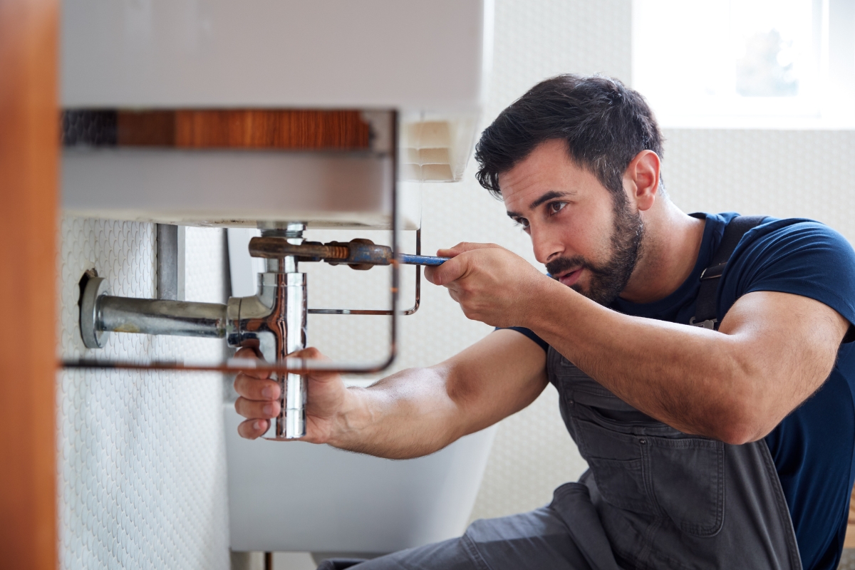 A man in work overalls is using a wrench to fix plumbing under a sink in a bathroom, demonstrating the precision and expertise often highlighted in content marketing for plumbers.