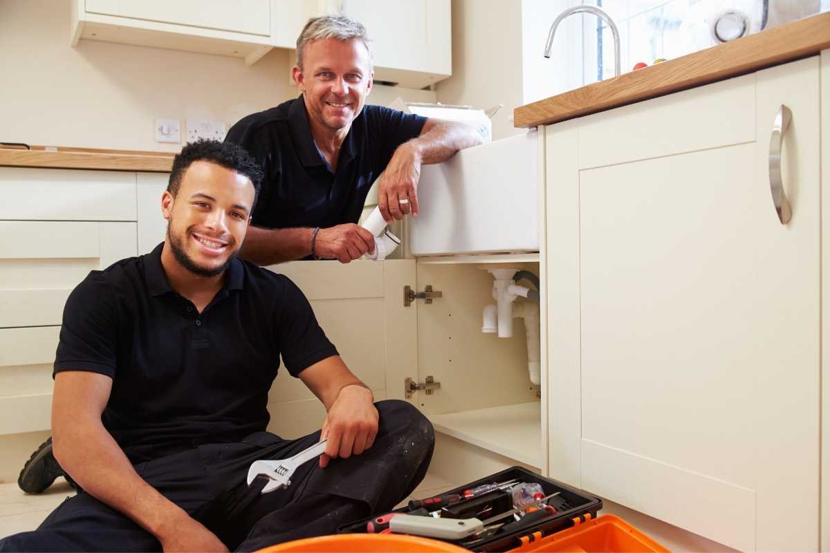 Two plumbers, one standing and one sitting, work on a sink in a kitchen setting. Various tools are visible on the floor and open cabinet. This scene could be ideal for content marketing for plumbers, showcasing their expertise and readiness to tackle any job.