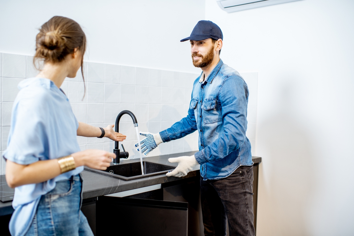 A plumber wearing a blue cap and gloves inspects a kitchen sink faucet while a woman stands nearby, gesturing towards the sink. It's moments like these that highlight the importance of content marketing for plumbers, showcasing their expertise and reliability in everyday scenarios.