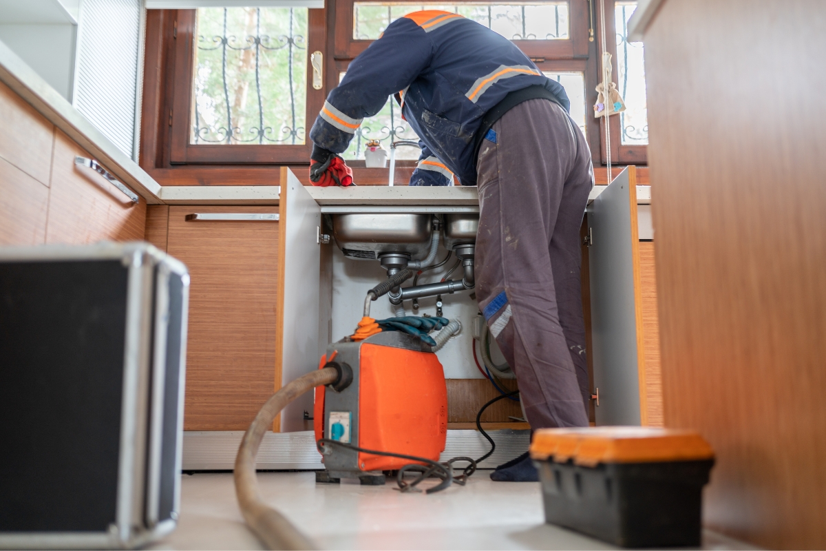 A plumber in a blue uniform is diligently working under a kitchen sink with a pipe cleaning machine and tools nearby, exemplifying excellent service that could be highlighted through effective content marketing for plumbers.