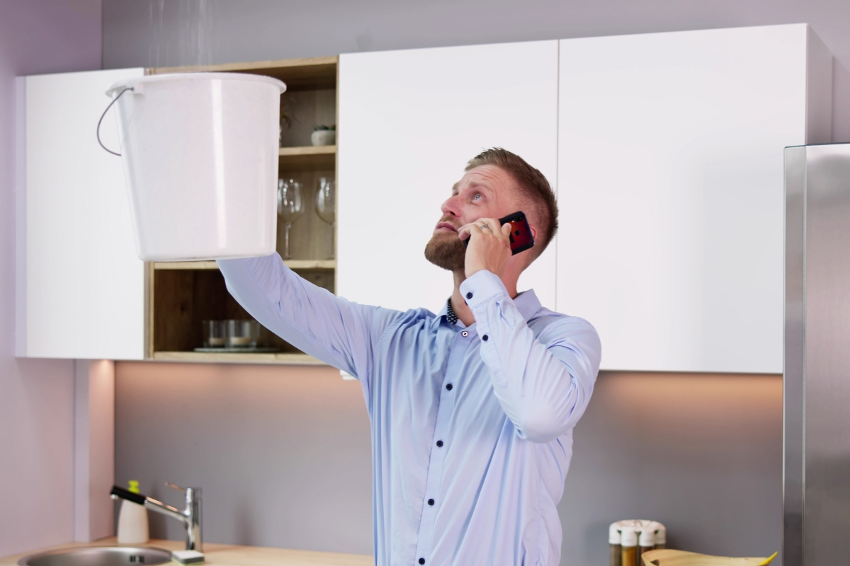 A man holds a white bucket to catch water leaking from the ceiling while talking on a phone in a modern kitchen, seemingly discussing content marketing for plumbers.