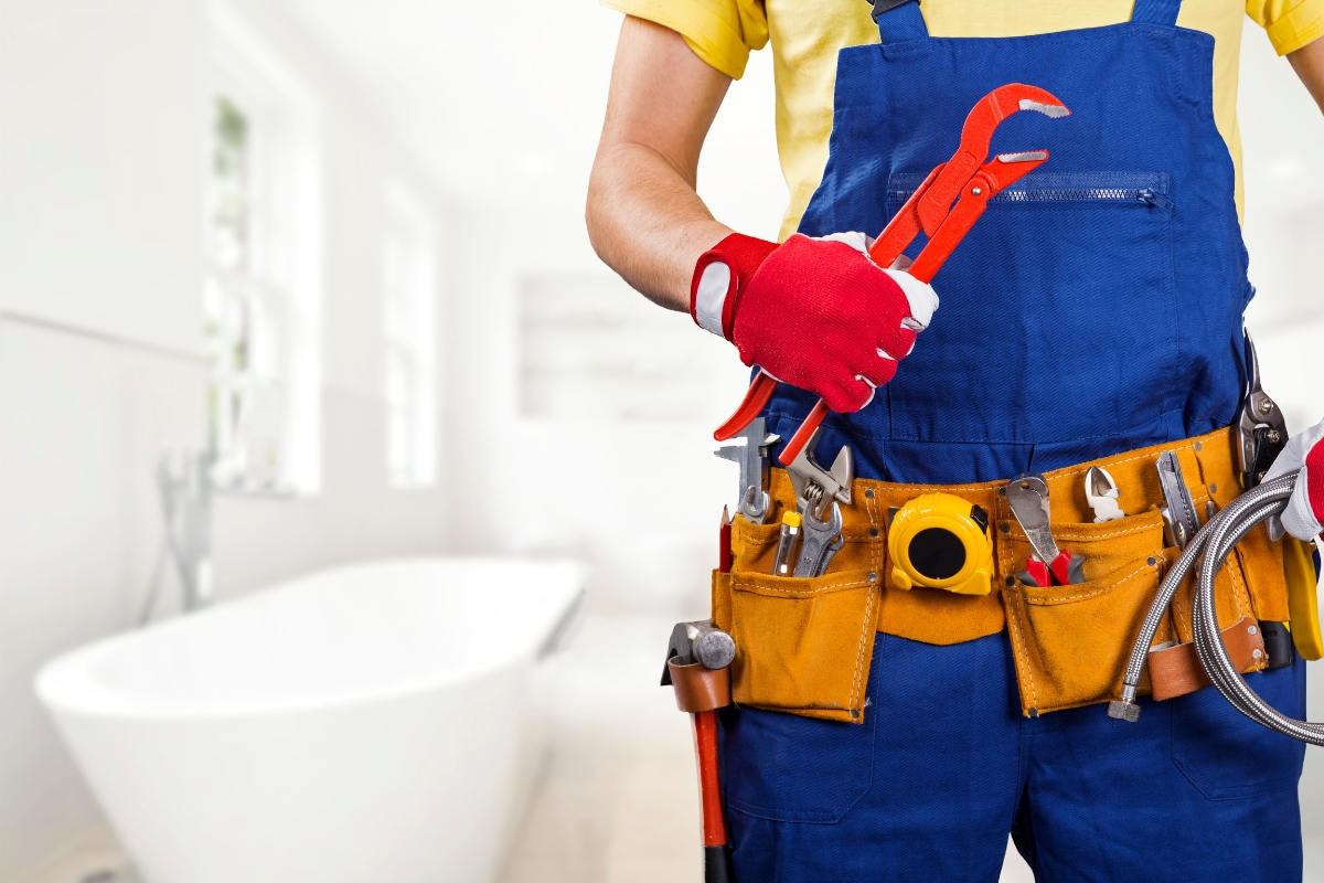 A plumber wearing a blue coverall and yellow shirt holds a pipe wrench. His tool belt brims with various tools like a tape measure, pliers, and screwdrivers. A modern bathroom is visible in the background, perfectly setting the scene for effective content marketing for plumbers.