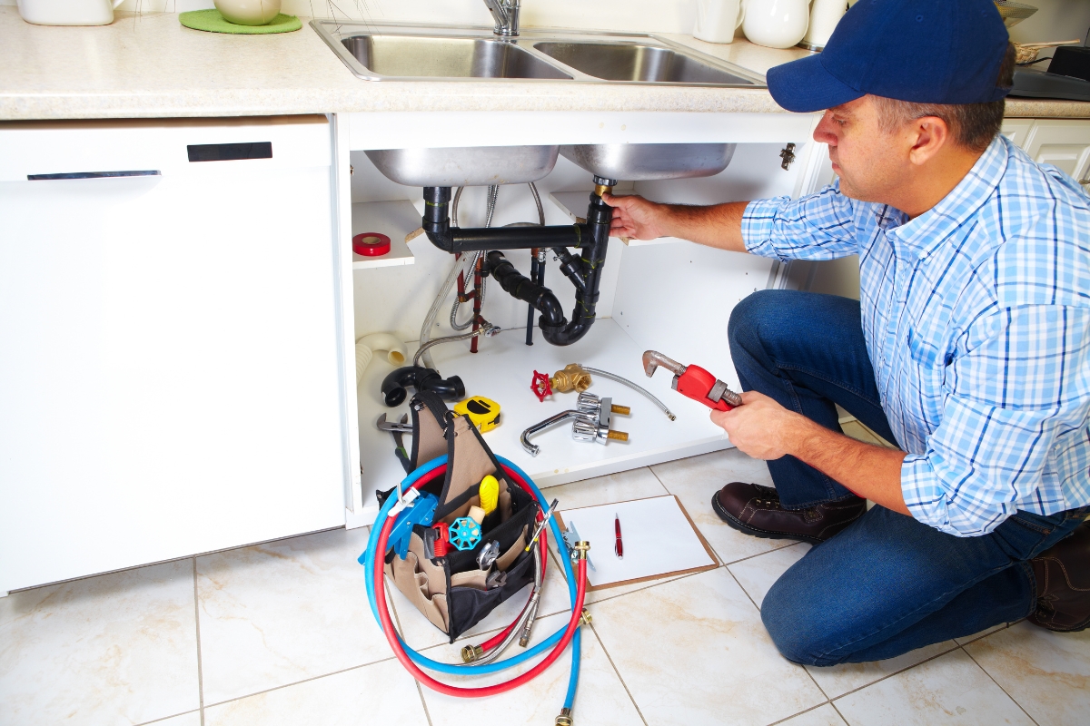 A plumber in a blue cap is kneeling under a kitchen sink, fixing the plumbing. Tools and a tool bag are on the floor next to him, showcasing his expertise—a perfect snapshot for content marketing for plumbers.