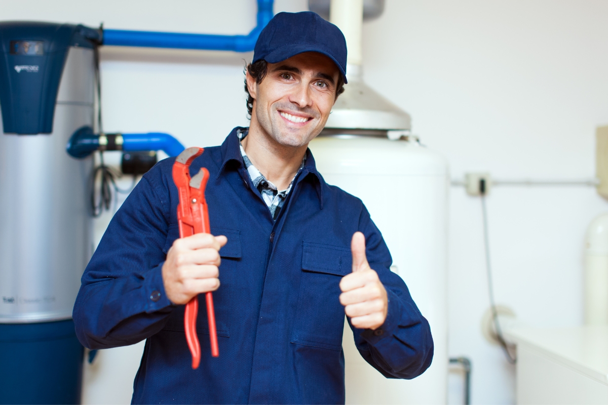 A technician in a blue uniform and cap holds a red pipe wrench in one hand and gives a thumbs-up with the other, standing in a room filled with various pipes and equipment, showcasing the reliability essential for email marketing for plumbers.