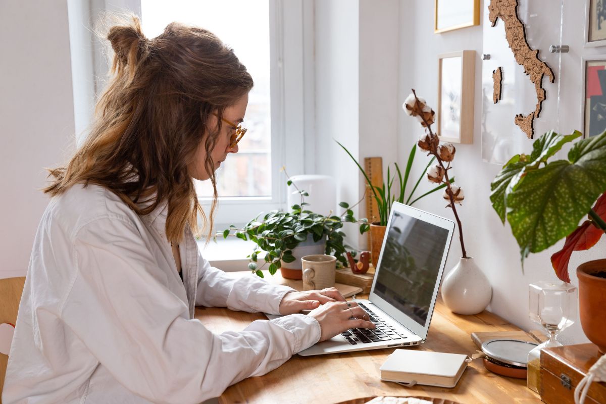 A woman with glasses and a white shirt sits at a wooden desk typing on a laptop, researching how to avoid AI detection. Plants, notebooks, and decorations surround her workspace.