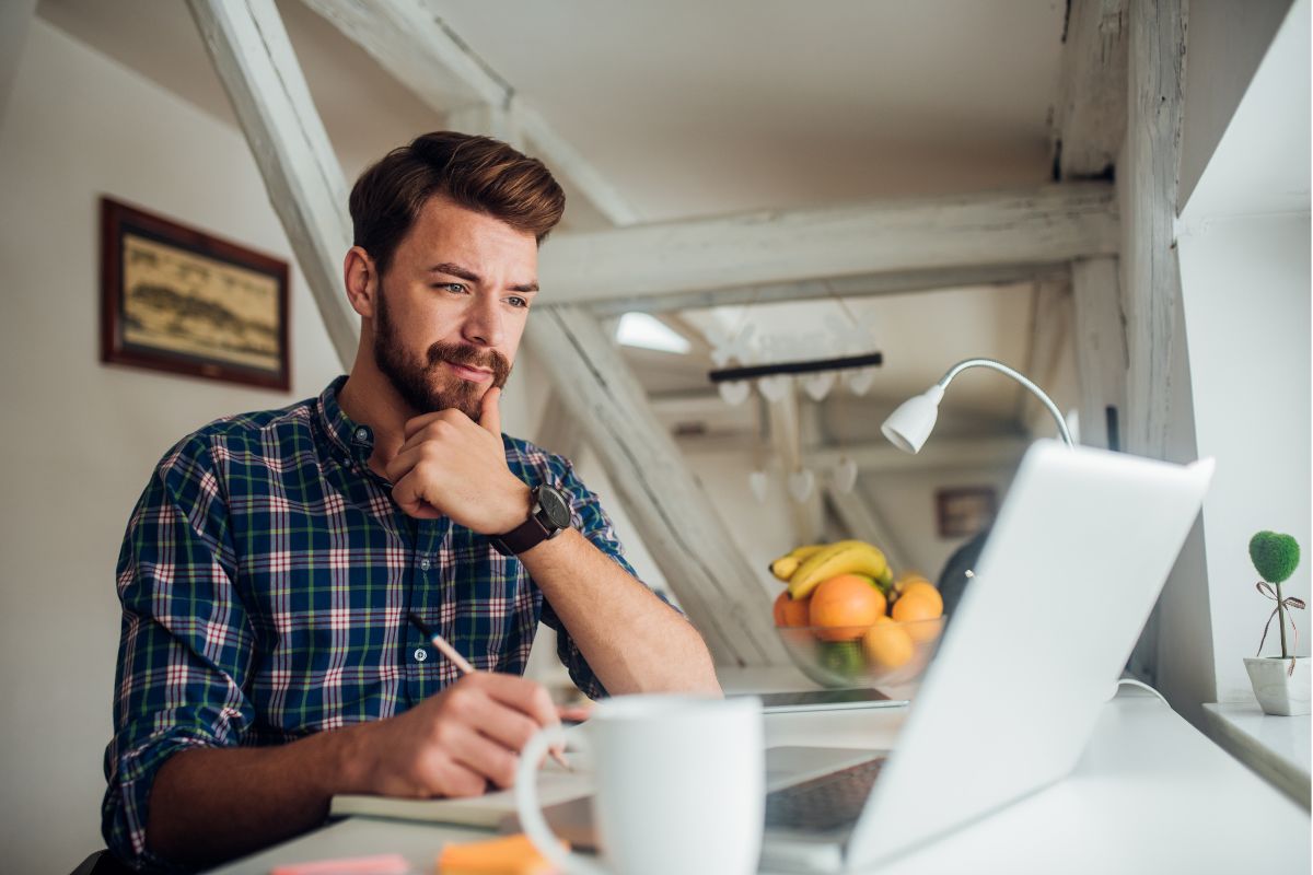 A man in a plaid shirt sits at a desk, focused on a laptop with his hand on his chin. Surrounding him are a coffee mug, papers, and a bowl of fruit as he researches how to avoid AI detection.