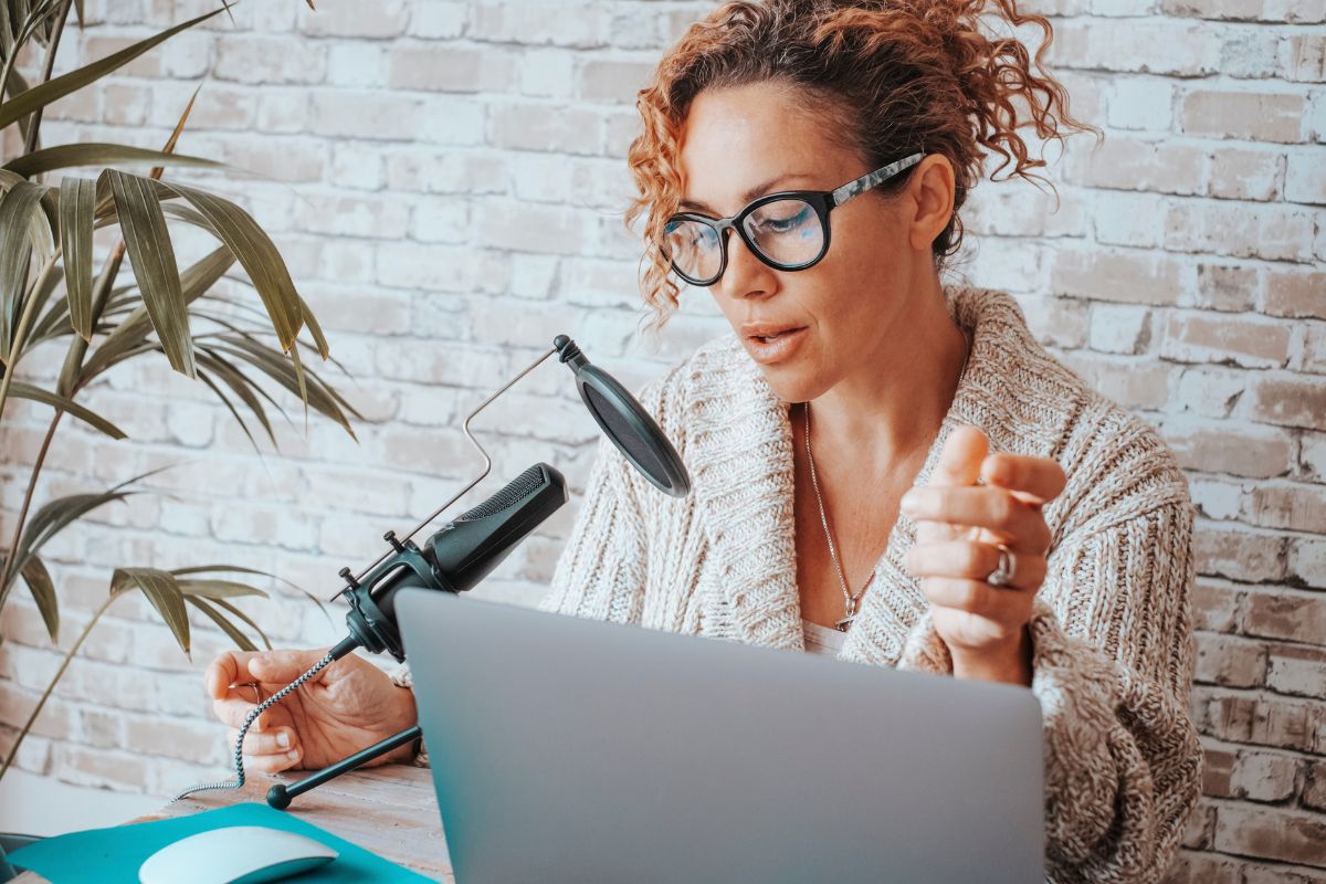 A person with curly hair and glasses is speaking into a microphone while using a laptop at a desk, likely discussing how to avoid AI detection, with a brick wall in the background. A potted plant is visible on the side.